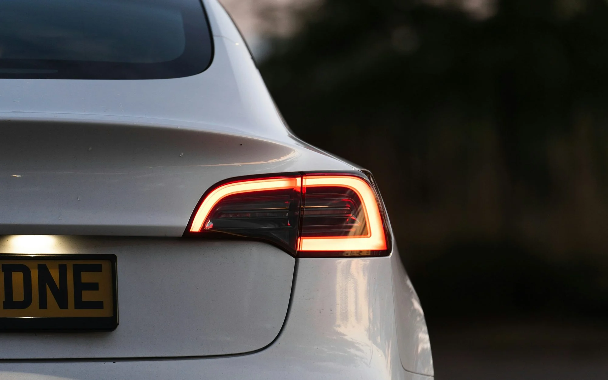 Rear of a white Tesla Model 3 at dusk with its tail light lit and a yellow UK number plate